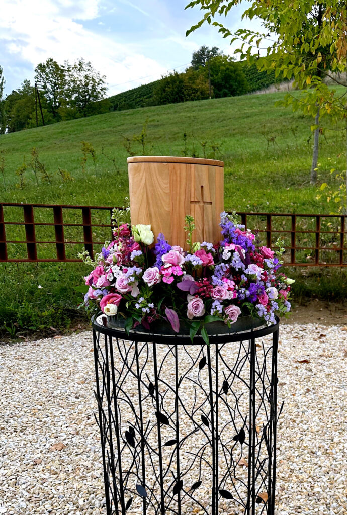 Urne aus Holz mit Blumenschmuck rundherum steht im Freien, dahinter grüner Hügel